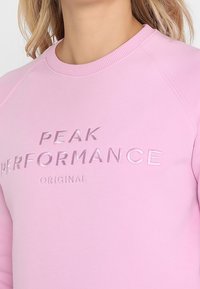 Růžová mikina s kulatým výstřihem a raglánovými rukávy. Na přední straně je ražený nápis: "PEAK PERFORMANCE ORIGINAL". Měkká textilie.