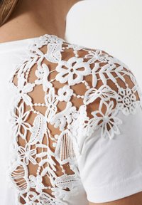 Camiseta blanca con intrincados detalles de encaje blanco con patrones florales en el hombro, que expone un fondo del tono de piel.