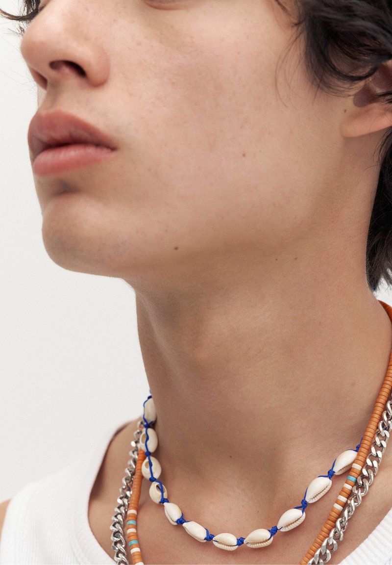Gros plan sur la partie inférieure du visage et le cou d'une personne portant trois colliers superposés : un collier coquillage bleu et blanc, un collier de perles orange et une chaîne en argent.