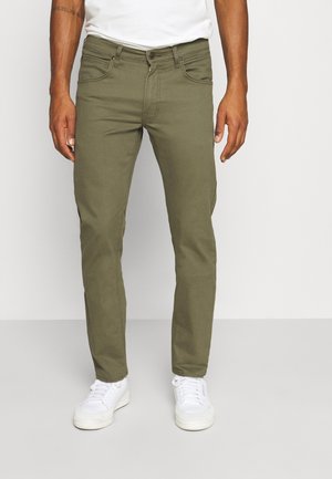 Man die olijfgroene slim-fit broek en witte sneakers draagt, staand tegen een eenvoudige lichte achtergrond.