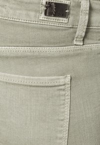Los pantalones de mezclilla verde claro presentan un bolsillo delantero con una costura visible. Una etiqueta de metal con un logo de caballo está unida a la cinturilla.