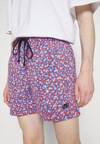 Person trägt blaue Shorts mit roten und orangefarbenen Punkten und schwarzem Nike-Logo, kombiniert mit einem schlichten weißen T-Shirt.