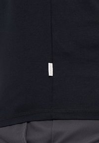 Tricou din bumbac negru, cu o mică etichetă albă a brandului pe partea inferioară, având o textură netedă și o croială standard.