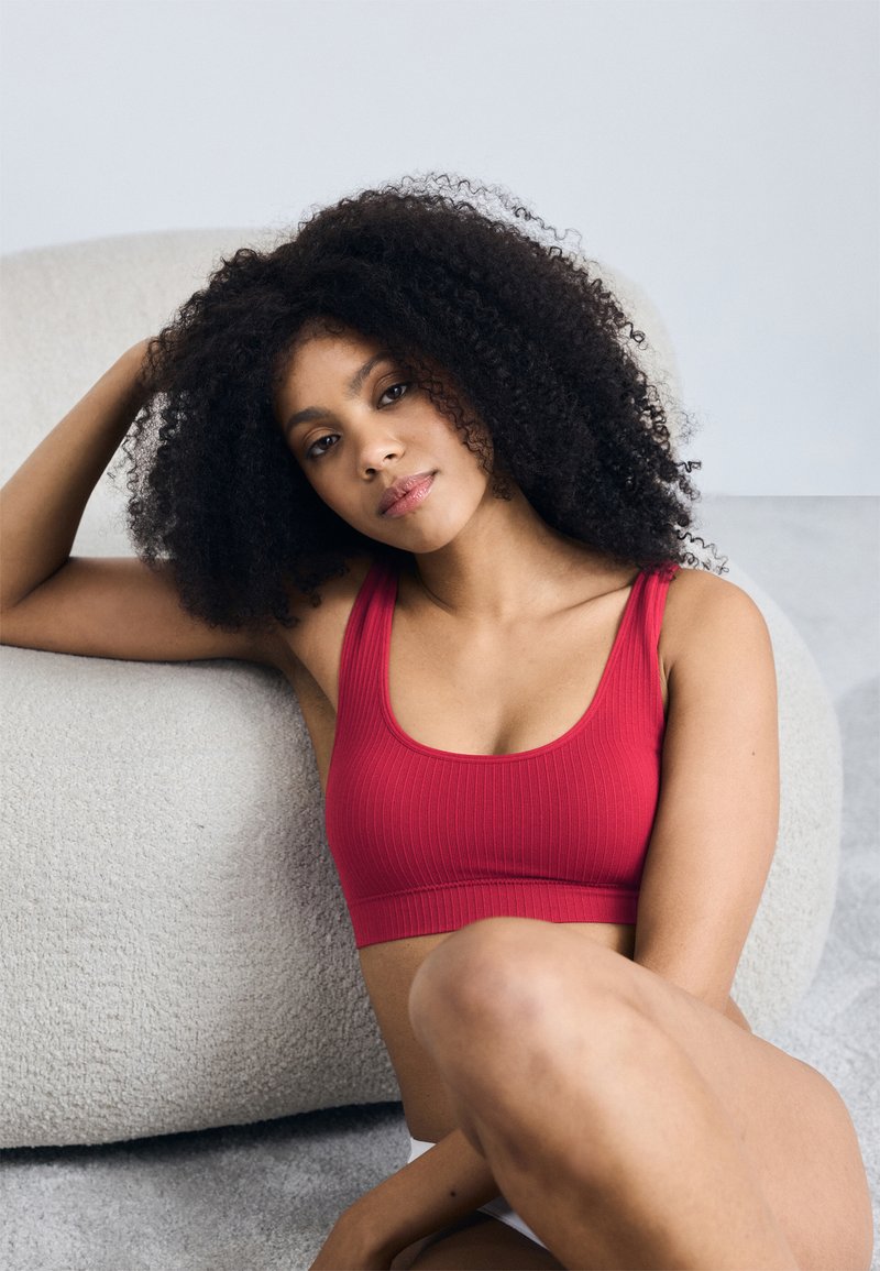 Červená športová podprsenka s širokými ramienkami a hlbokým výstrihom. Model sedení na textúrovanom svetlom pozadí.