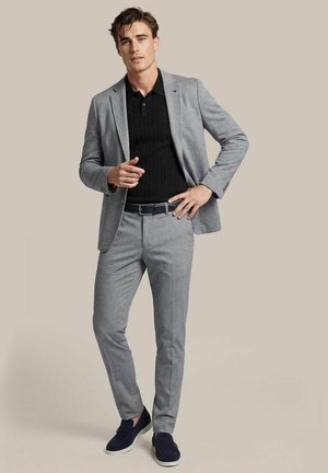 Man in lichtgrijs pak en zwart overhemd, staande met de ene hand op de heup en de andere vooruit wijzend tegen een neutrale achtergrond.