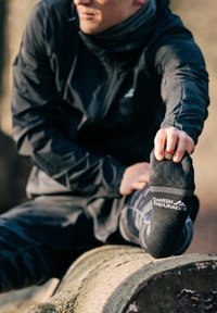 Schwarze Sportsocke mit "DANISH ENDURANCE"-Logo, strukturierter Oberfläche und gestreiften Akzenten. Getragen von einer Person, die im Freien auf einem Baumstamm dehnt.