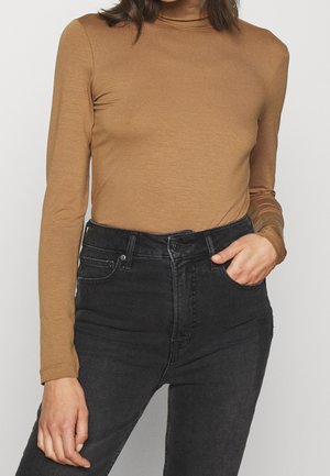 Vrouw draagt een aansluitende bruine coltrui met lange mouwen, gestopt in een zwarte hooggetailleerde spijkerbroek, met één hand in de zak.