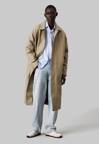 Beige lange jas, lichtblauw overhemd met knopen, lichtblauwe jeans en crèmekleurige sneakers. Glad materiaal, ontspannen pasvorm en eenvoudig ontwerp.