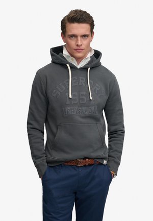 Ung mand iført en mørkegrå Superdry hættetrøje med hvide snører, en hvid skjorte med krave indenunder, blå bukser og et brunt flettet bælte.