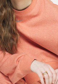 Coral sweatshirt with a crew neck, long sleeves, and a subtle silver Nike logo on the chest. Smooth texture and relaxed fit.