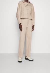 Veste en jean beige avec fermeture à boutons et design cropped, associée à un pantalon droit assorti. Chaussures blanches à semelle épaisse.