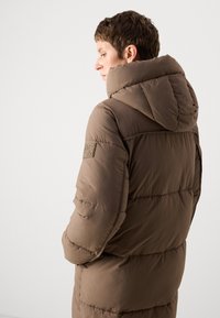Personne portant un manteau d'hiver rembourré marron avec une grande capuche et un patch logo sur la manche, tourné sur le côté devant un fond uni.