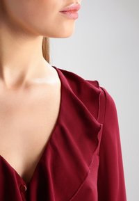 Volto e collo di una donna che indossa una blusa bordeaux con collo a volant su uno sfondo chiaro e semplice.