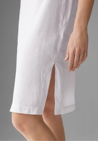 Abito bianco realizzato in un tessuto leggero, con una spaccatura laterale e un orlo dritto. Una mano visibile tocca delicatamente il tessuto vicino alla spaccatura.