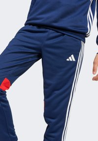 Marineblaue Trainingshosen mit weißen Seitenstreifen und roten Akzenten, die ein schlankes Design, eine glatte Textur und ein Adidas-Logo aufweisen.