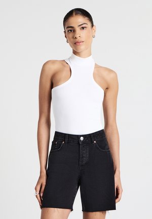 Donna che indossa un top smanicato bianco asimmetrico con collo alto e pantaloni shorts in denim neri a vita alta con orlo grezzo, in piedi davanti a uno sfondo bianco.