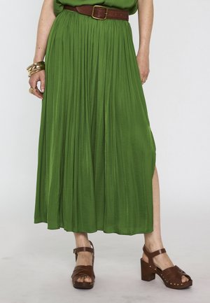 Femme portant une jupe midi plissée verte avec une ceinture marron, des sandales marron à talons, ainsi que des bracelets et bagues en or aux mains.