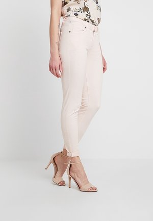 Femme portant un jean skinny rose clair, un top fleuri et des sandales à talons hauts beige avec bride à la cheville, debout sur fond blanc.