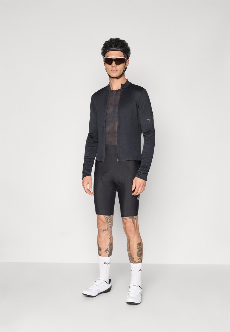 Schwarzes langärmliges Radtrikot mit Reißverschluss vorne, getragen über einem schwarzen kurzärmeligen Unterhemd und kombiniert mit schwarzen gepolsterten Shorts und weißen Radschuhen.