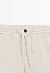 Light beige linen shorts with an elastic waistband, drawstring, and a black button closure. Smooth texture and simple design.