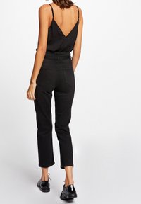 Kvinna klädd i en svart ärmlös topp med djup V-rygg, högmidjade svarta byxor och svarta glänsande snörskor som står mot vit bakgrund.