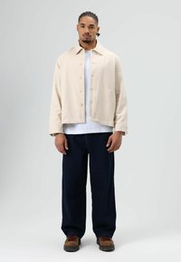 Beige cropped jack met knoopsluiting, gecombineerd met een wit T-shirt en donkerblauwe jeans met wijde pijpen. Bruine veter schoenen. Eenvoudig ontwerp.