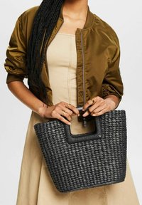 Borsa tote intrecciata nera con finitura testurizzata, caratterizzata da una maniglia quadrata e un'apertura spaziosa, abbinata a una giacca bomber marrone.