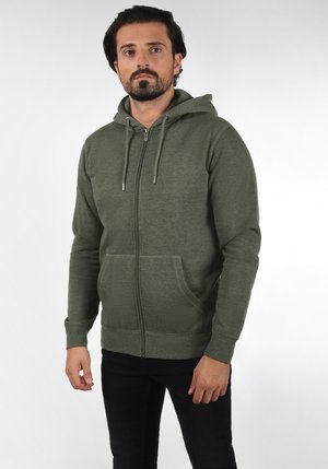 Sudadera con cremallera en verde oliva de tejido suave con un acabado texturizado, que cuenta con un bolsillo frontal y capucha con cordón. Llevada con jeans negros.