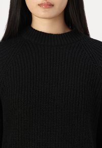 Maglione nero lavorato a maglia con una texture a coste, scollatura rotonda e maniche raglan. Il tessuto appare spesso e caldo.