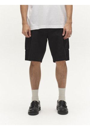 Osoba oblečená v čiernych cargo šortkách, bielych ponožkách, čiernych kožených topánkach a bielej tričku, stojaca na jednoliatom bielom pozadí.