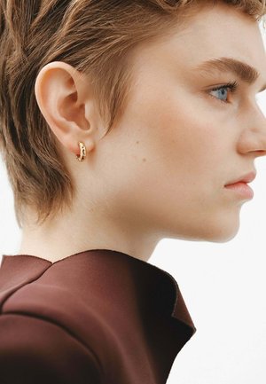 Pendiente de aro dorado con un acabado texturizado, llevado en la oreja izquierda de una persona con cabello corto castaño, contra un fondo neutro.