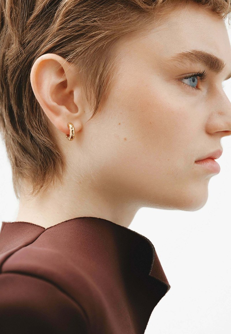 Pendiente de aro dorado con un acabado texturizado, llevado en la oreja izquierda de una persona con cabello corto castaño, contra un fondo neutro.