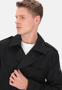 Sort trenchcoat med struktureret overflade, stor krave og knapdekorationer. Modellen holder kraven og har en hvid skjorte under.
