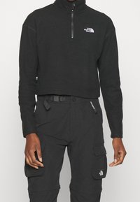 Personne portant une polaire noire North Face à demi-fermeture éclair et un pantalon cargo noir avec des écussons de marque et une boucle de ceinture réglable visible.