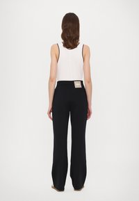 Black wide-leg pants with a high waistband, featuring a small label on the back. The model wears a white tank top. Smooth fabric texture.