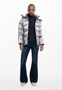 Silverfärgad puffjacka med hög krage, bärs över en marinrandig tröja och utsvängda mörka jeans. Vita sneakers fulländar outfiten.
