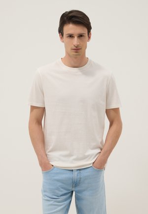 Jeune homme debout, les mains dans les poches, portant un t-shirt blanc uni et un jean bleu clair sur un fond neutre.