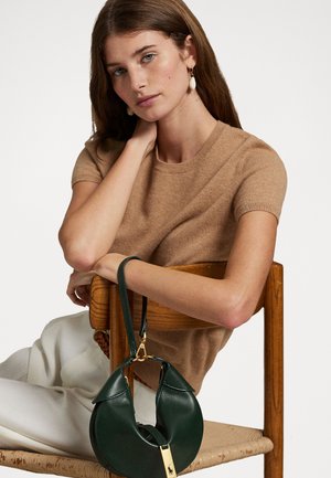 Un pull marron à manches courtes associé à un sac en cuir vert avec un design incurvé et des détails en métal doré, posé sur une chaise en bois.