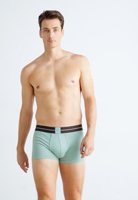 Boxeri scurți de culoare verde mentă cu un tiv negru, având un buzunar frontal și un design fără cusături, realizati dintr-un material elastic și moale.