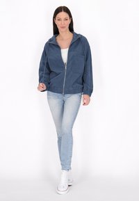 Denim jack, blauw, met ritssluiting aan de voorkant, verstelbare capuchon, twee voorkeurs, elastische manchetten en zoom, gepaard met lichtblauwe jeans en witte sneakers.