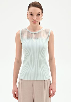 Giovane donna che indossa un top azzurro senza maniche a costine con strisce verticali trasparenti al collo e pantaloni beige su uno sfondo neutro.