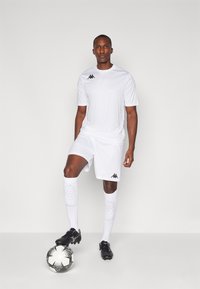 Balts futbola tērps ar īsām piedurknēm un šortiem, kuram ir melns logs. Modelis stāv ar melnu sporta apavu uz melnbalta futbola bumbas.