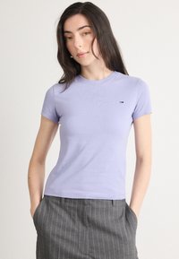 Kortærmet lilla t-shirt med rund halsudskæring og en lille logo-detalje. Stof ser blødt og tætsiddende ud, parret med grå stribede bukser.