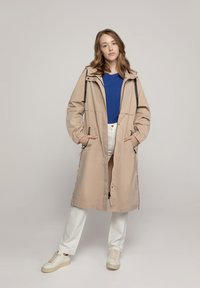 Beige waterdichte parka met een kraag, verstelbare manchetten en twee voorkruiszakken, gedragen over een blauwe top, gecombineerd met witte broeken en sneakers.