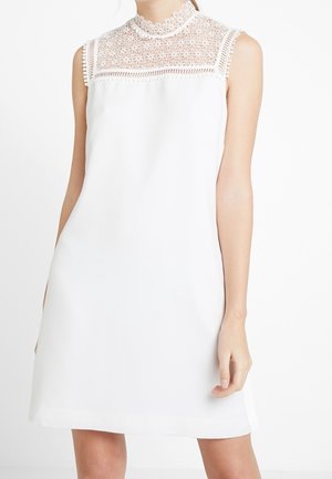 Ærmeløs hvid kjole med høj blondehalsudskæring og blomsterblonde detaljer hen over brystet, båret af en stående person.