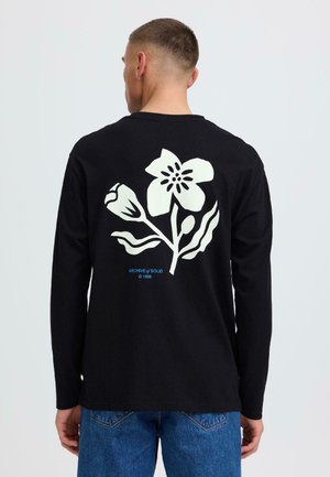 Mann trägt schwarzes Langarmshirt mit großem weißem Blumenmotiv und blauem Text "ARCHIVE of SOLID © 1999" auf dem Rücken, kombiniert mit blauen Jeans.