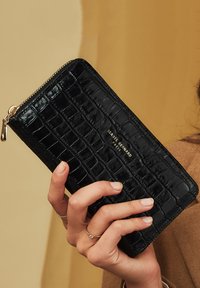 Schwarze Geldbörse aus Leder mit Krokodilmuster und goldenem Reißverschluss sowie Branding, von einer Hand gehalten, mit glatter, glänzender Textur.