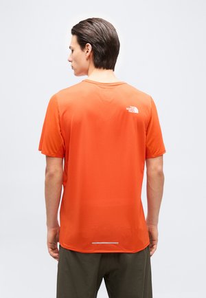 Mann mit dunkelbraunen Haaren, der ein oranges The North Face T-Shirt mit kurzen Ärmeln und dunkle Hosen trägt, von hinten vor einfarbigem Hintergrund.