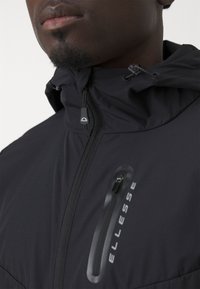 Schwarze Kapuzenjacke mit einer Reißverschlusstasche, verfügt über ein mattes Finish und ein Logo auf der Vorderseite. Glatte Textur mit verstellbaren Kordeln.