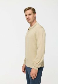 Polo de culoare bej, cu mânecă lungă, guler cu două butoane și manșete cu nervuri, realizat dintr-un material neted. Purtați-l cu pantaloni bleumarin.